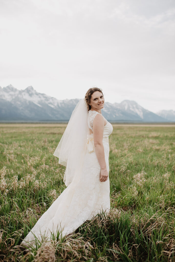 jackson hole photographers photographs bride in a field looking over her shoulder with the tetons behind her for her wyoming elopement