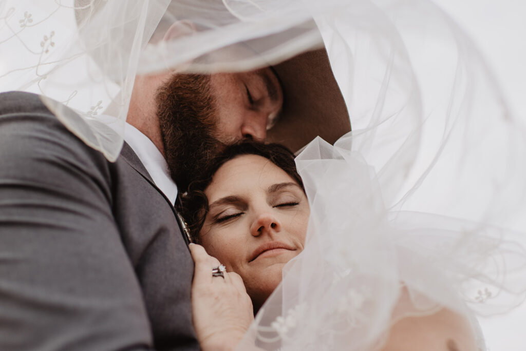 groom hugging his bride as she rests her head on his chest and he leans down to kiss her head while they stand under her veil captured by jackson hole photographers