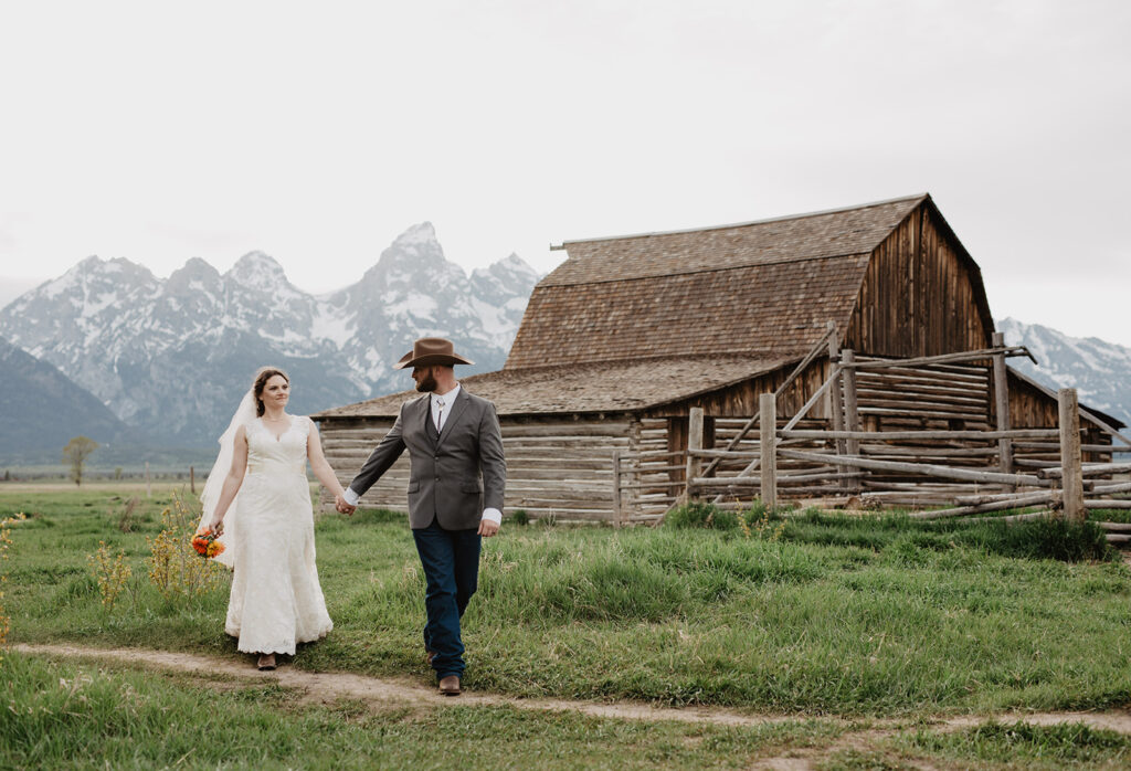 Grand Teton wedding with bride and groom holding hands and walking in front of a barn at Mormon Row with the Tetons in the distance captured by jackson hole photographers