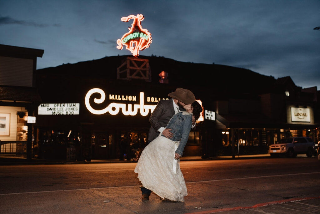 cowboy bar in Jackson with bride and groom outside of the bar with the bride being dipped by her groom in the road in front of the bar at night captured by jackson hole photographers for wyoming elopement