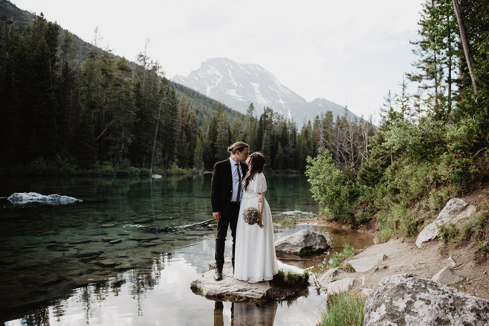 lake Jackson Hole wedding with bride and groom standing on a rock in a lake with a forest and the Tetons in the distance captured by Jackson Hole elopement photographer