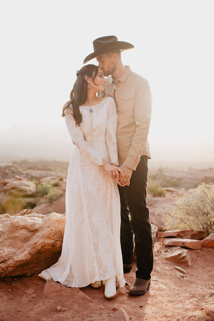bride and groom standing on top of a red rock cliff for their Moab elopement holding hands with the groom standing behind his bride and kissing her on the head captured by utah elopement photographer 