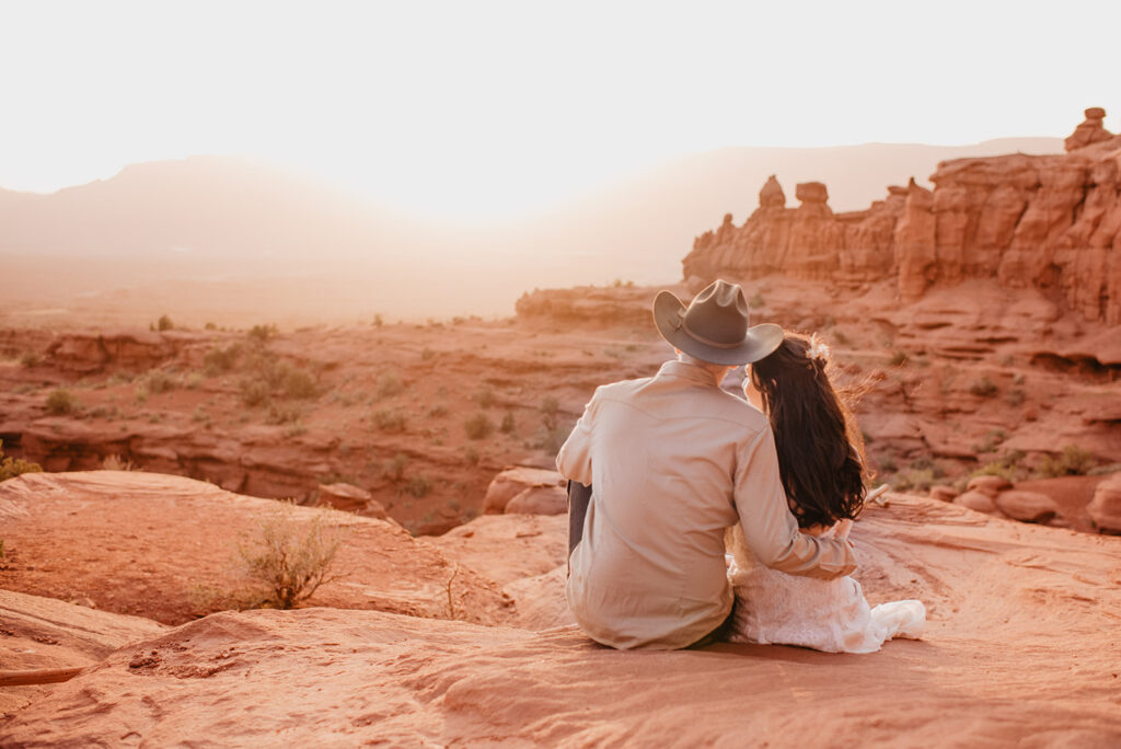 arches national park wedding with bride and groom sitting together on top of a red rock cliff and watching the sun set in the distance as they cuddle together