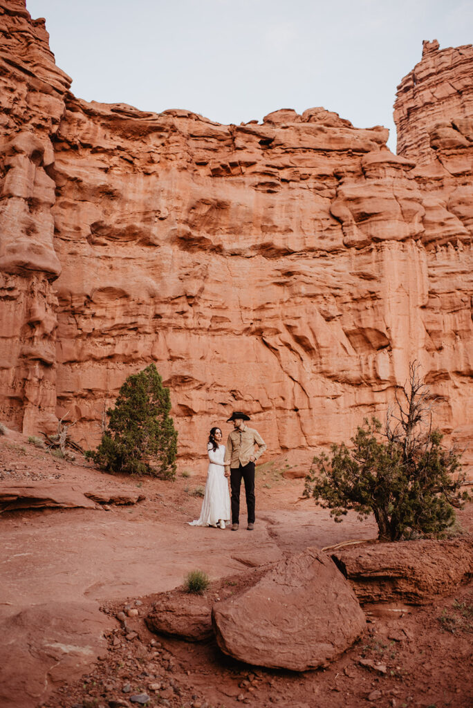 utah elopement photographer captures unique wedding pictures with bride and groom standing together and holding hands while looking to one another with a larhe red rock cliff behind them