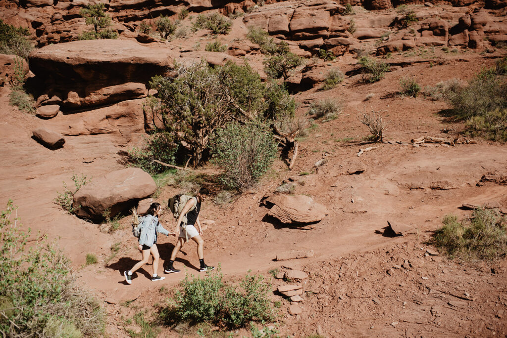 Moab elopement with bride and groom hiking to their ceremony location with Utah elopement photographer as the couple holds hands and walk up a red rock hill 