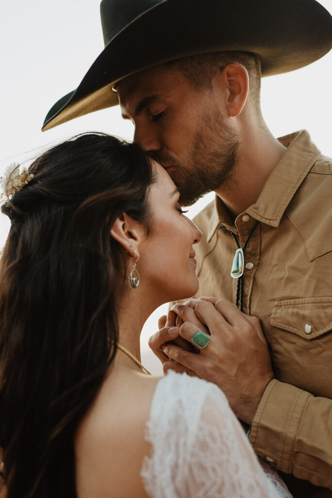 cowboy groom kisses his brides forehead while holding her hands to his chest during their Moab elopement with Utah elopement photographer