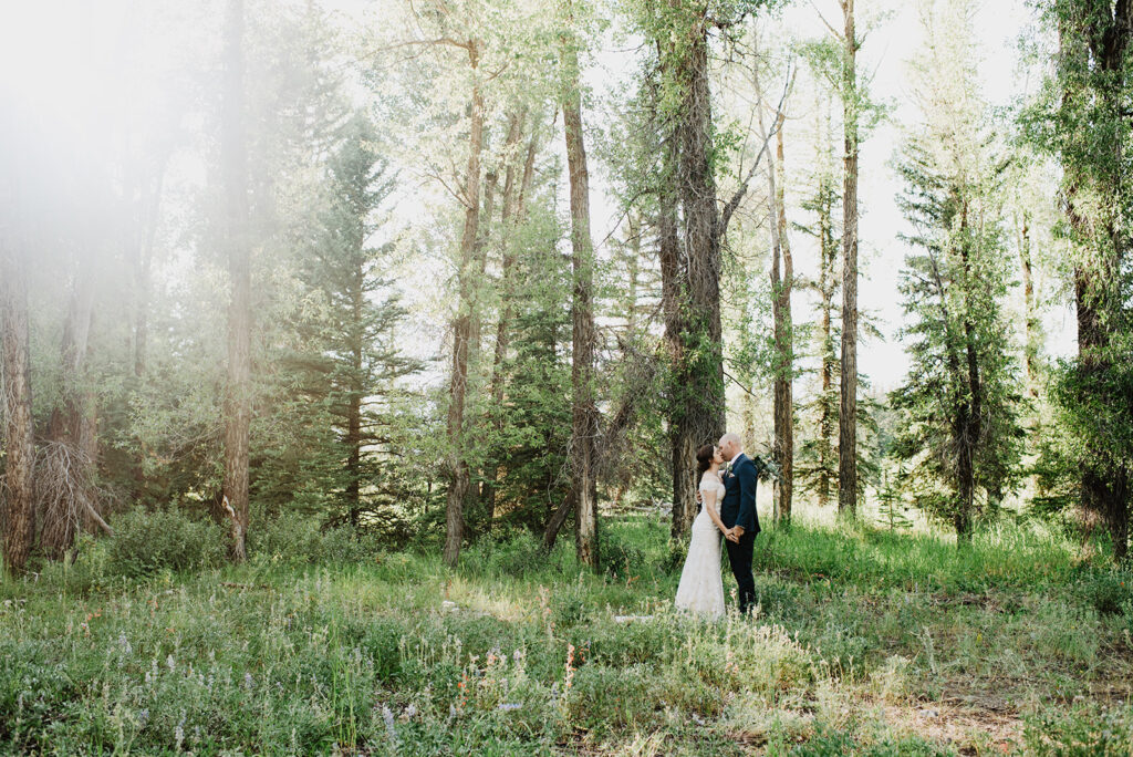 woodland wedding with bride and groom holding hands and kissing in the woods of the tetons with the sun shining through the trees with wildflowers surrounding them for their Jackson Hole wedding