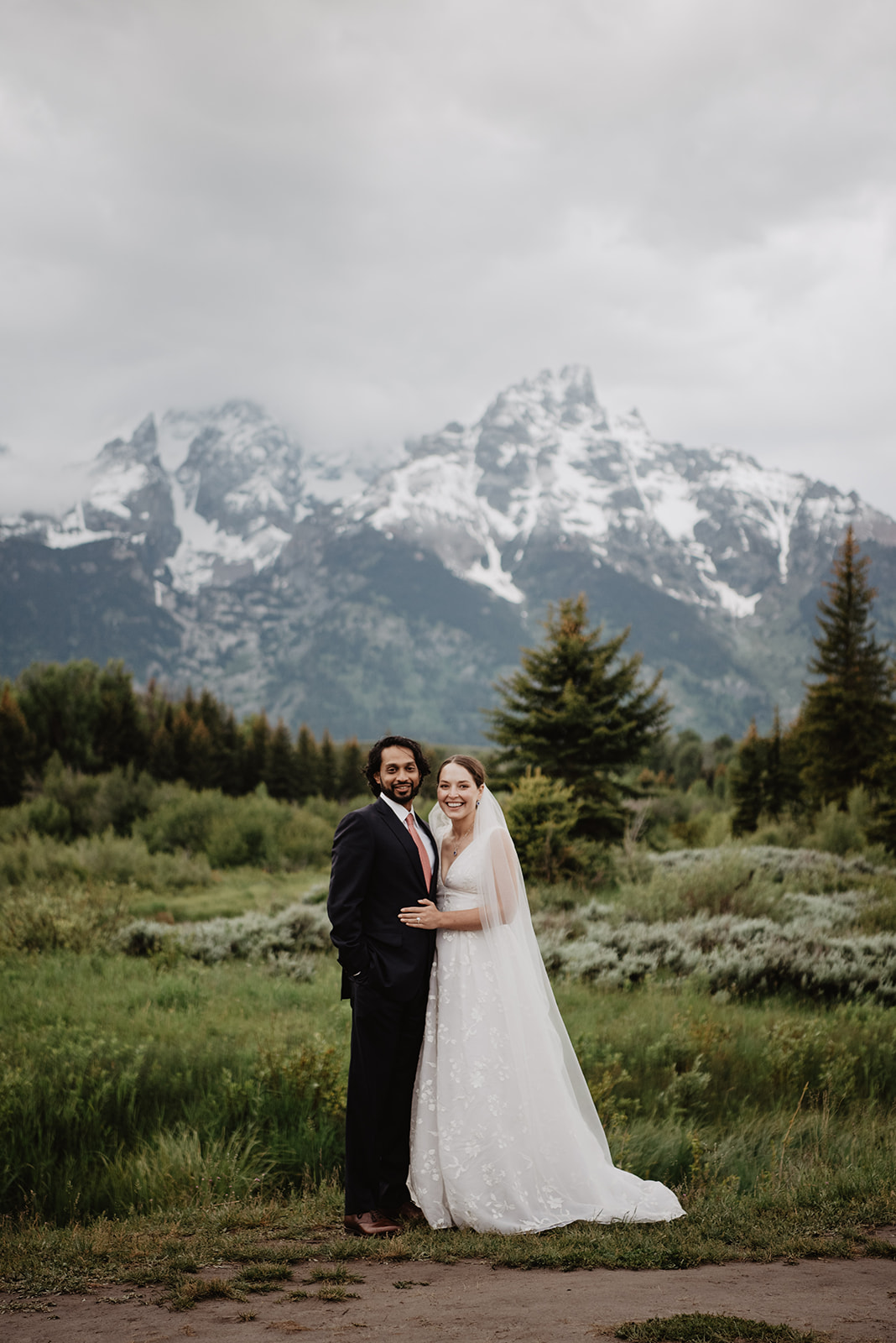 bride and groom standing together in a field with the tetons covered in snow behind them as they smile together for their Jackson Hole wedding