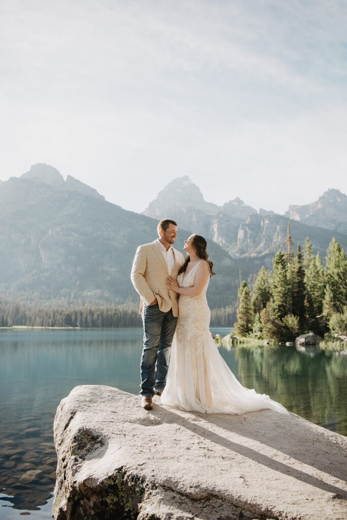 bride standing with her groom and resting her hand on his chest as they stand on a boulder next to the lake with the grand tetons in the distance 