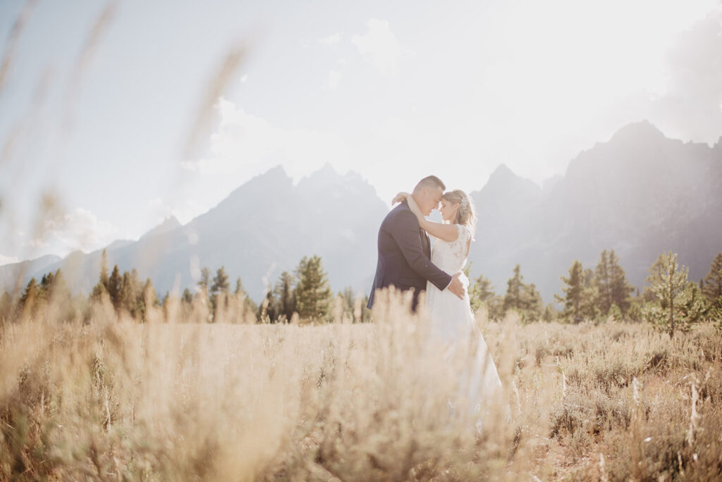 jackson hold photographer capture bride and groom embracing in a field with the sun setting over the mountains with tall grass surrounding the couple for their grand teton wedding 