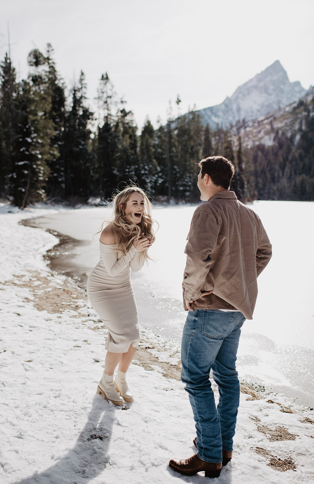 A woman beams with joy and excitement on a snowy lakeshore as she faces her partner, with the rugged Tetons and evergreen trees framing the background. This candid moment, full of laughter and love, captures the essence of a memorable engagement in Jackson Hole, ideal for couples seeking an elopement photographer who appreciates the natural beauty of Wyoming's landscapes.