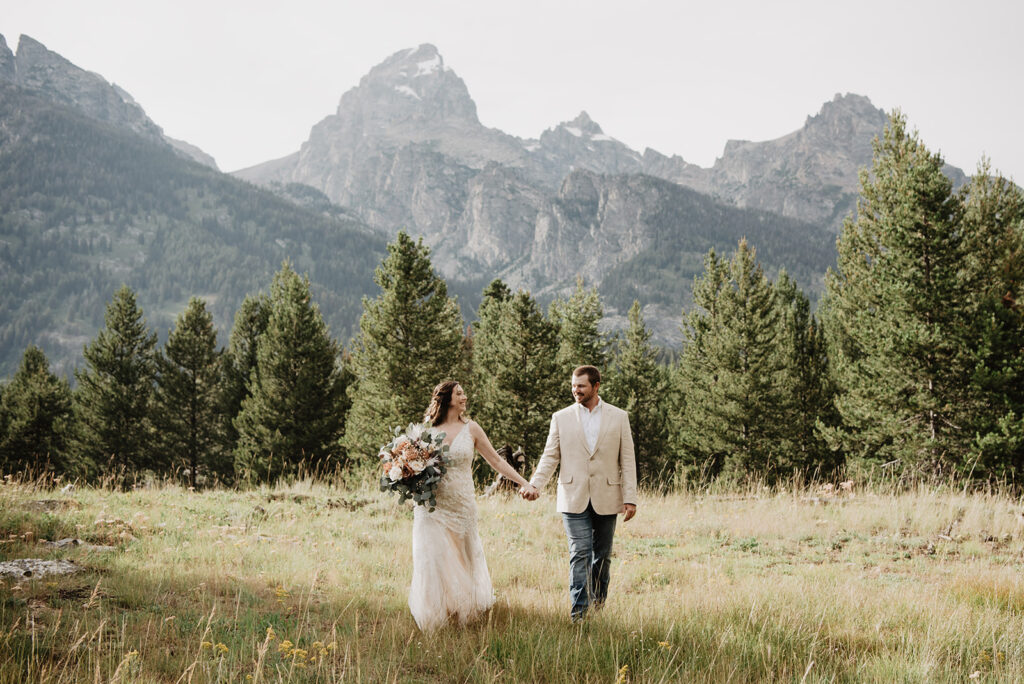 Grand Teton wedding picture with bride and groom holding hands and walking through a field with the trees in the distance and mountains right behind the trees