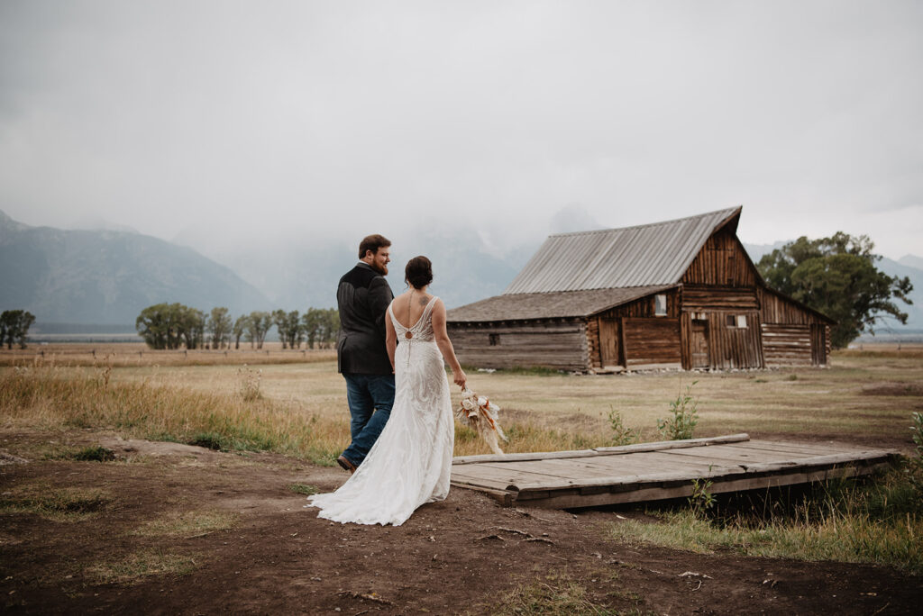 Jackson Hole photographers capture bride and groom holding hands and walking over a small bridge that covers a creek as they walk towards a barn with a line of trees in the distance with fog covering the mountains