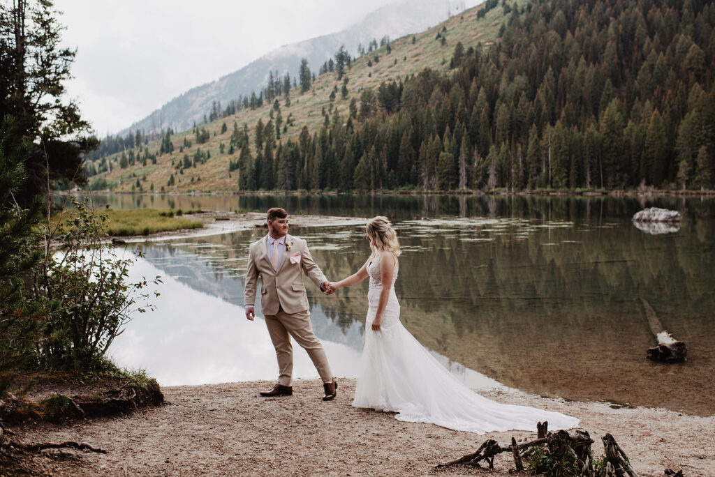 Jackson Hole Wedding Photographer captures groom leading bride along lake in Grand Teton National Park