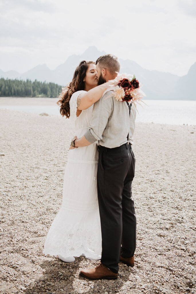 Grand teton elopement with bride holding her arms around the grooms neck while holding her bouquet and kissing while on a beach in the tetons