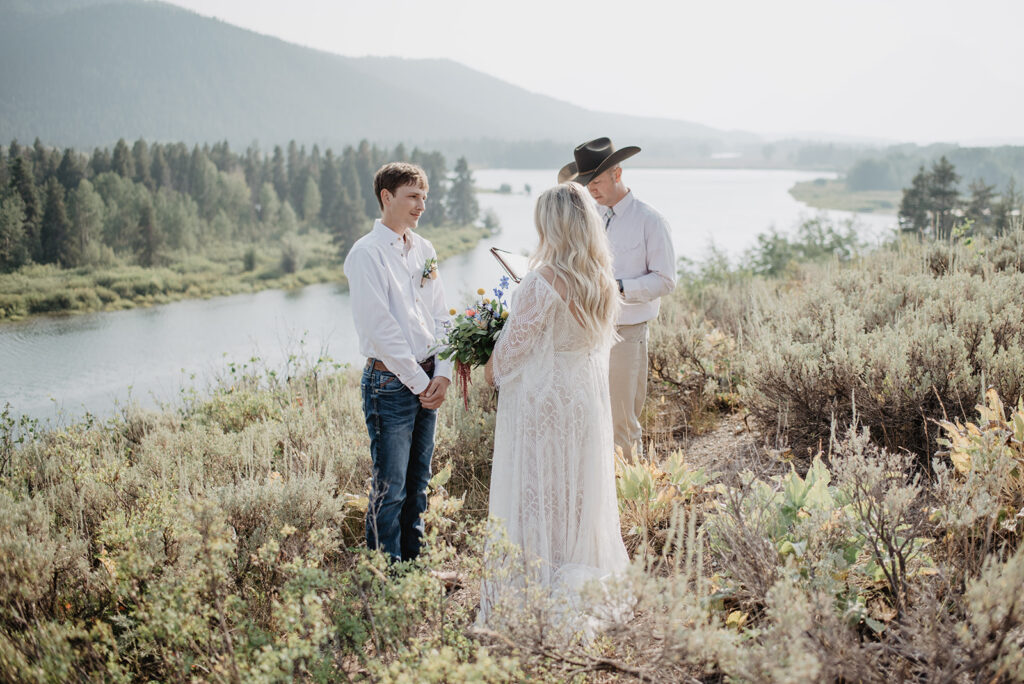 man and woman standing with their officiant at Oxbow Bend over the snake river in the distance with the sun shining and reflecting over the water