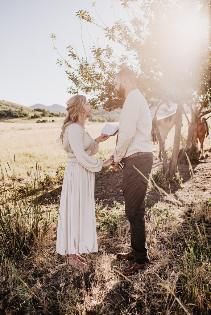 bride reading her vows from a vow book with the sun setting through a tree that two horses are tied to