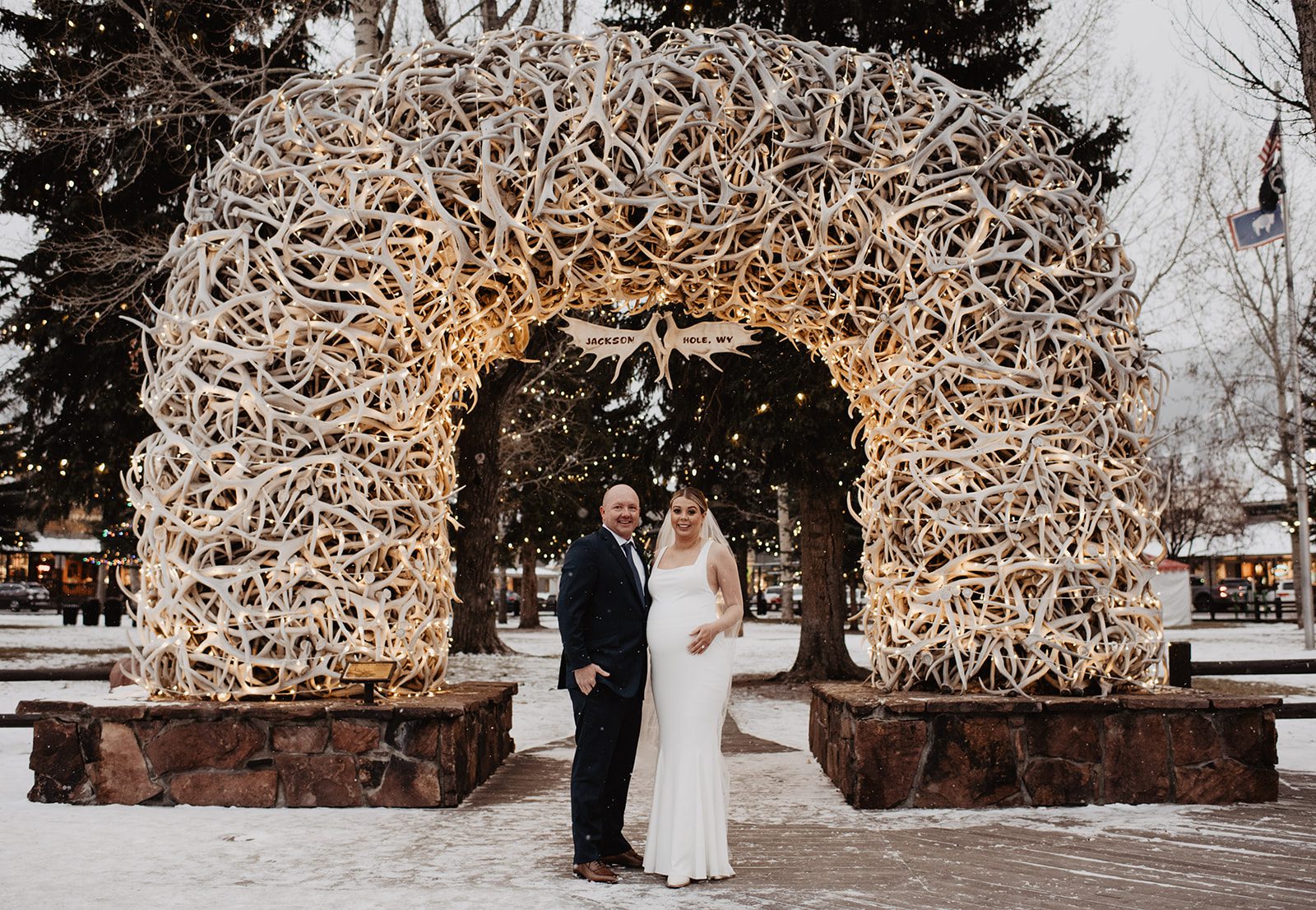 A bride and groom stand together beneath the iconic illuminated antler arch in Jackson Hole, WY, surrounded by gently falling snow and twinkling lights. The bride, in a sleek white gown with a flowing veil, smiles alongside her groom, dressed in a dark suit. The archway, adorned with numerous antlers and soft lights, creates a magical winter backdrop, capturing a memorable moment for couples celebrating with a Jackson Hole elopement photographer.