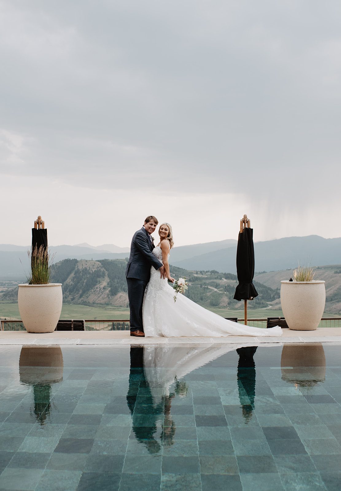 A bride and groom embracing by a reflective pool, with the dramatic landscape of the Grand Tetons and rolling hills as their backdrop, captured during their stunning Jackson Hole elopement at Amangani