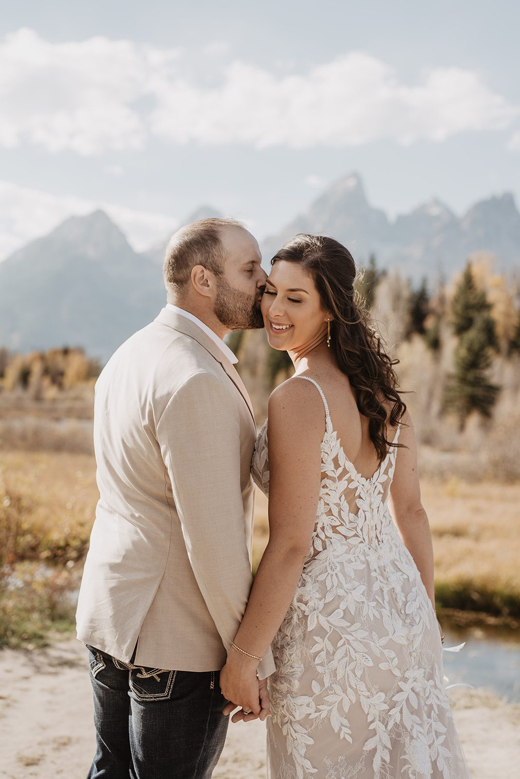 Jackson Hole photographer photographs bridal portrait of bride and groom holding hands and the groom kissing the brides cheek with a mountain backdrop