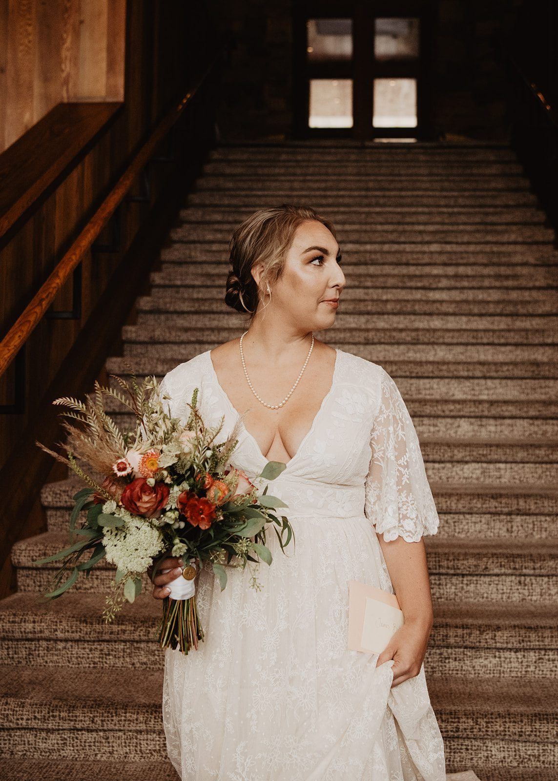 Bride holding a colorful bouquet and envelope, standing at the base of a grand staircase inside Amangani, wearing a lace wedding dress, captured by a Jackson Hole photographer for an Amangani wedding.