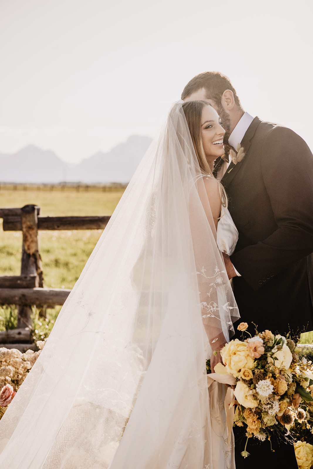 Close-up of a bride smiling while holding a yellow and white floral bouquet, with the groom leaning in affectionately during their Grand Teton elopement, captured by a Jackson Hole photographer against a rustic wooden fence and mountain backdrop.